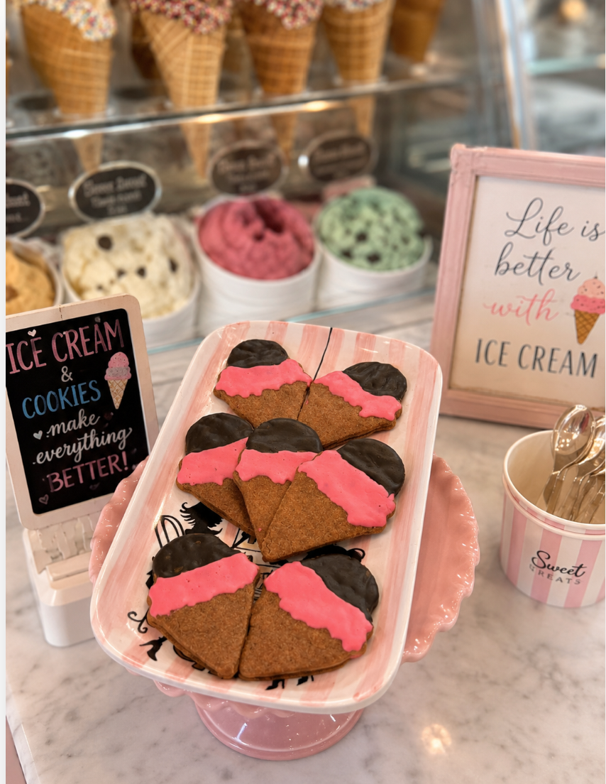 Ice cream cone-shaped dog treats with pink icing and chocolate-dipped tops displayed on a pink striped serving tray inside a pastel ice cream shop scene. The setup features marble counters, colorful ice cream tubs, waffle cones, decorative signs, and soft bakery-style lighting for a whimsical dessert shop aesthetic.