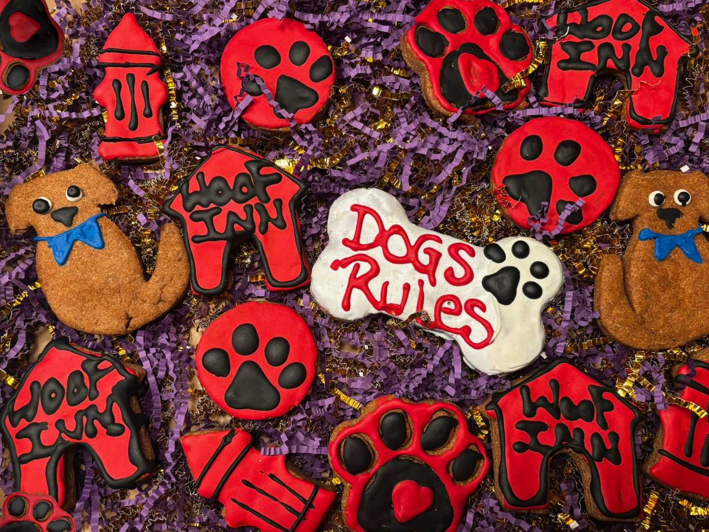 Assorted handmade organic dog cookies with red and black icing on decorative purple packaging, including paw prints, dog shapes, and “Dogs Rule” bone cookie