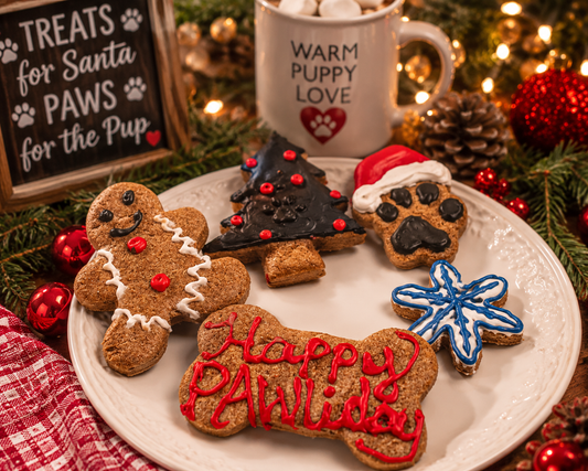 Festive handmade Christmas dog cookies displayed on a cozy holiday table surrounded by glowing Christmas lights, pinecones, evergreen branches, and red ornaments. The decorated organic cookies include a gingerbread pup, Christmas tree, snowflake, paw print with Santa hat, and a ‘Happy PAWlidays’ bone cookie arranged on a white plate beside a holiday mug and rustic Christmas sign